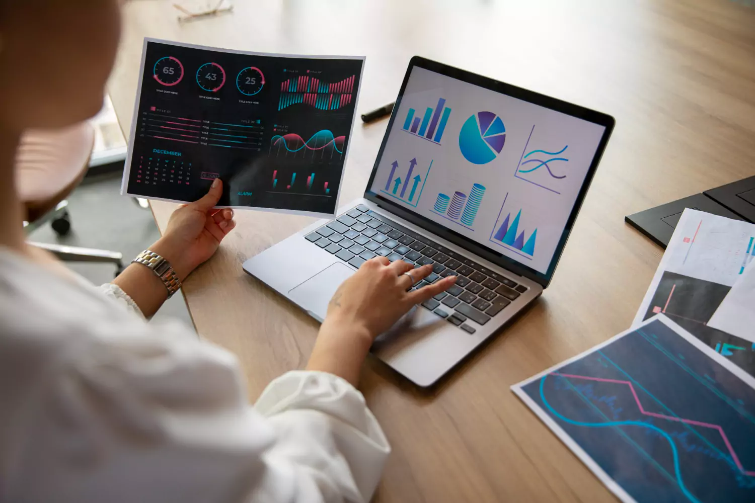 On the laptop screen and on paper, Amazon analytics reports are displayed as a woman works on them.