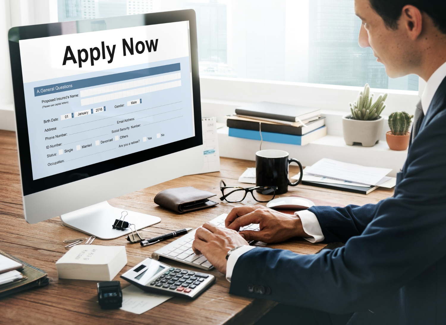 A man wearing black coat sitting on a table in front of a computer screen with some papers, keyboard and calculator. "Apply Now" is written on the screen of computer